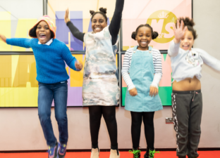 Four young, primary aged children jump for joy in front of a large colourful poster with the Ministry of Stories logo