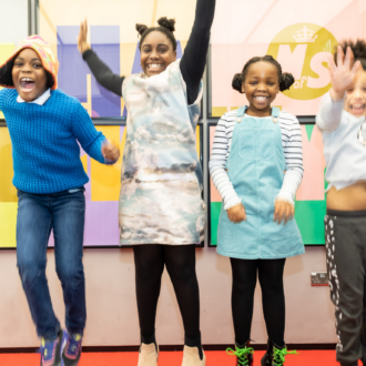 Four young, primary aged children jump for joy in front of a large colourful poster with the Ministry of Stories logo