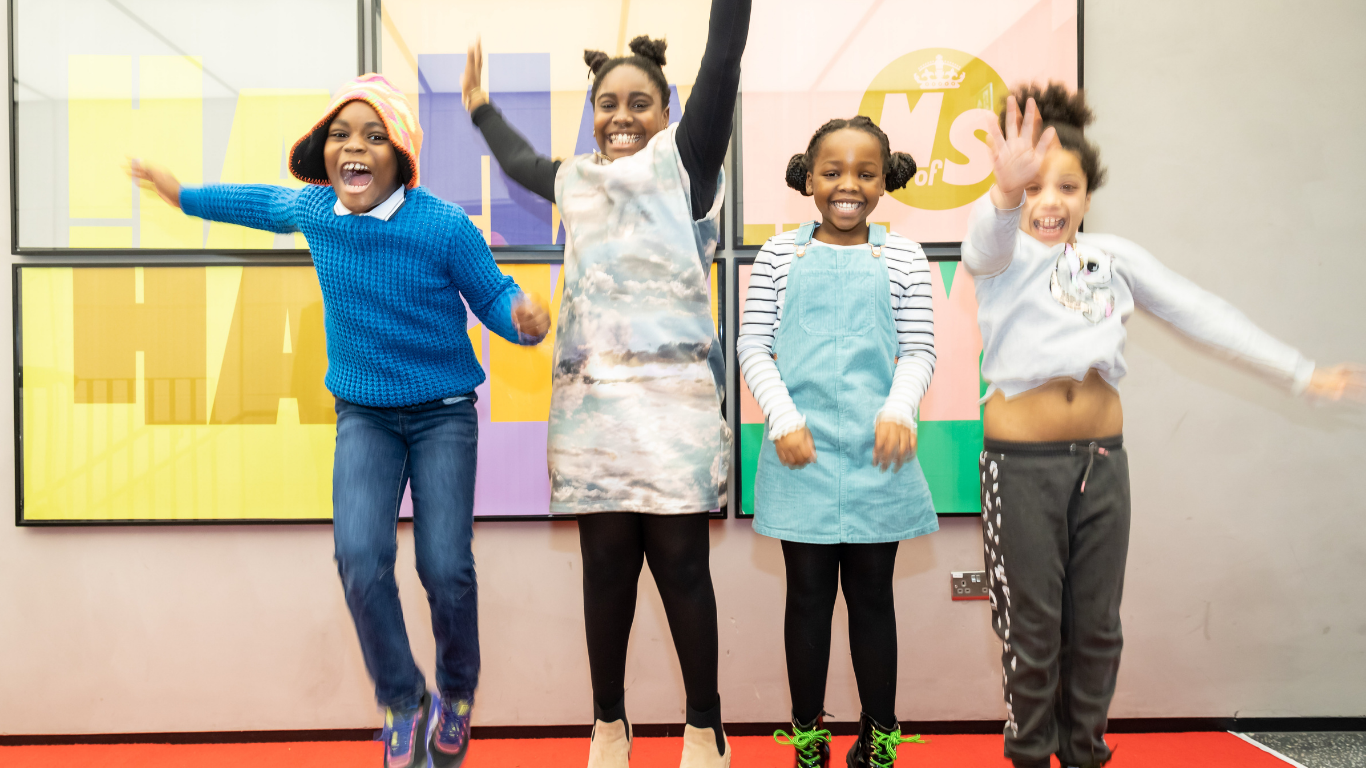 Four young, primary aged children jump for joy in front of a large colourful poster with the Ministry of Stories logo