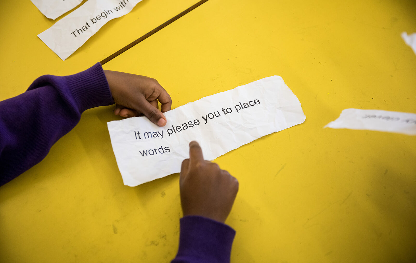 Two hands lay on a bright yellow table holding a large strip of paper that reads 'it may please you to place words...'