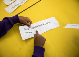 Two hands lay on a bright yellow table holding a large strip of paper that reads 'it may please you to place words...'