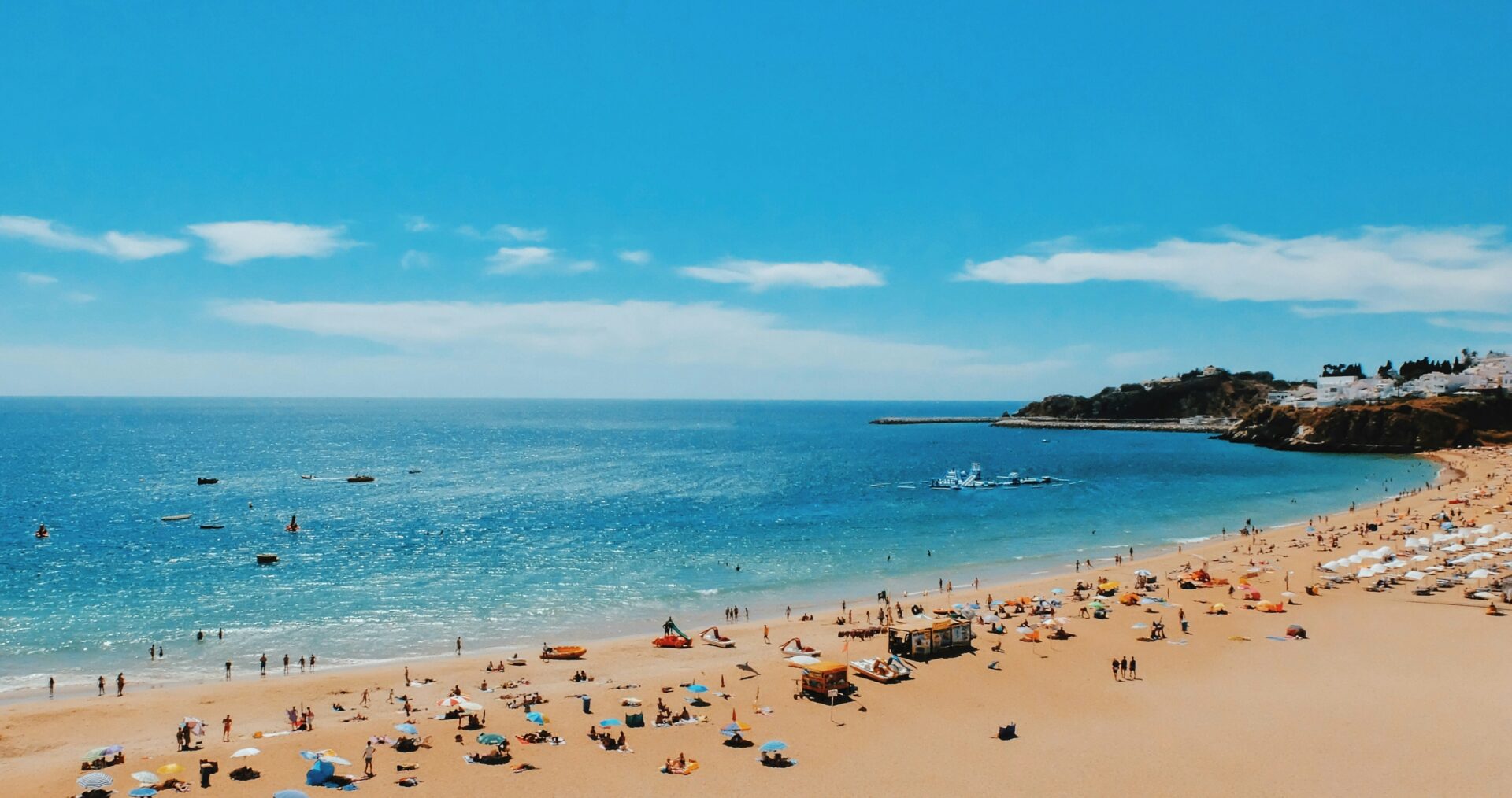 A landscape view of a golden sandy beach, bright blue sky and deep blue sea. There are colourful towels and umbrellas on the beach, and everything feels warm and peaceful.