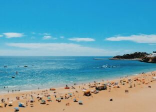 A landscape view of a golden sandy beach, bright blue sky and deep blue sea. There are colourful towels and umbrellas on the beach, and everything feels warm and peaceful.