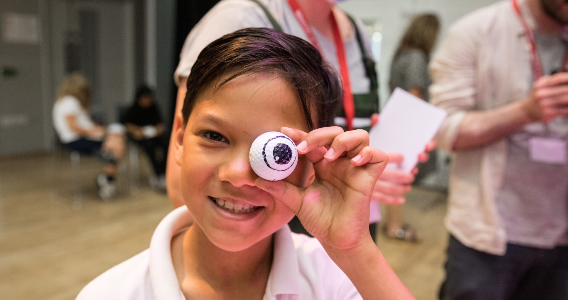 A young, primary ages boy with short dark hair grins at the camera. He holds a fake eye, that looks like a ping pong ball, up to his own eye. He wears a white polo shirt, with a school crest on the front.