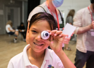 A young, primary ages boy with short dark hair grins at the camera. He holds a fake eye, that looks like a ping pong ball, up to his own eye. He wears a white polo shirt, with a school crest on the front.