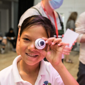 A young, primary ages boy with short dark hair grins at the camera. He holds a fake eye, that looks like a ping pong ball, up to his own eye. He wears a white polo shirt, with a school crest on the front.