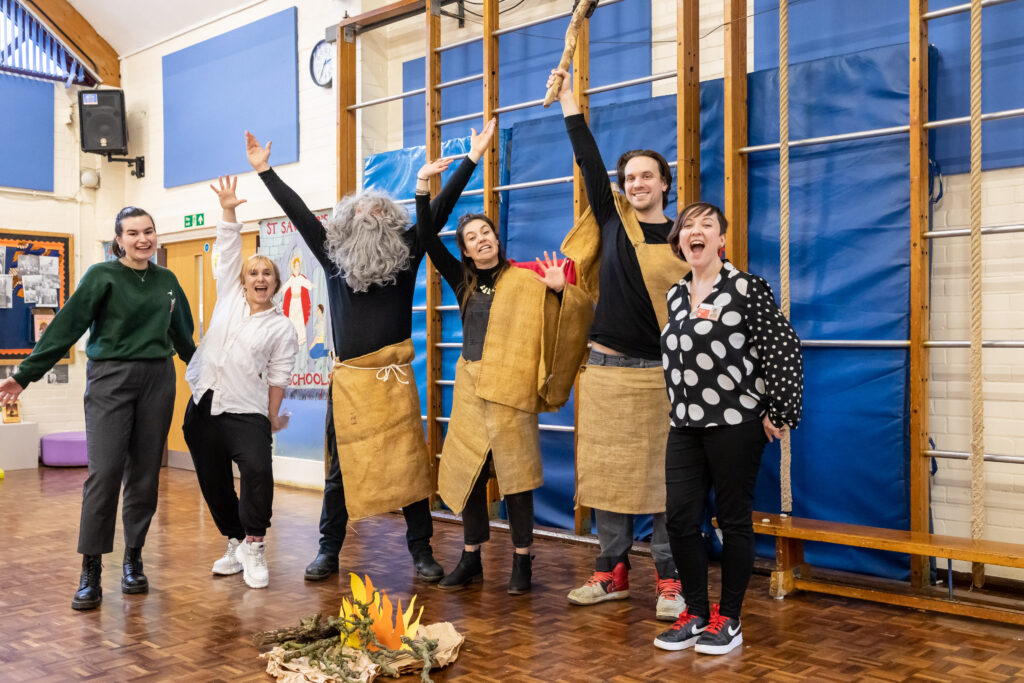 A group of adults - dressed up as stone age people, with large fake beards and brown loin cloths over black clothes - stand grouped in a school gymnasium, smiling and grining at each other, posing in a playful way.