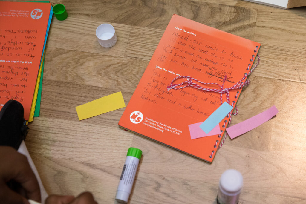 A orange book lays on a wooden floor. It is surrounding by colourful paper and glue sticks. The book is covered in a child's writing and looks inviting and fun.