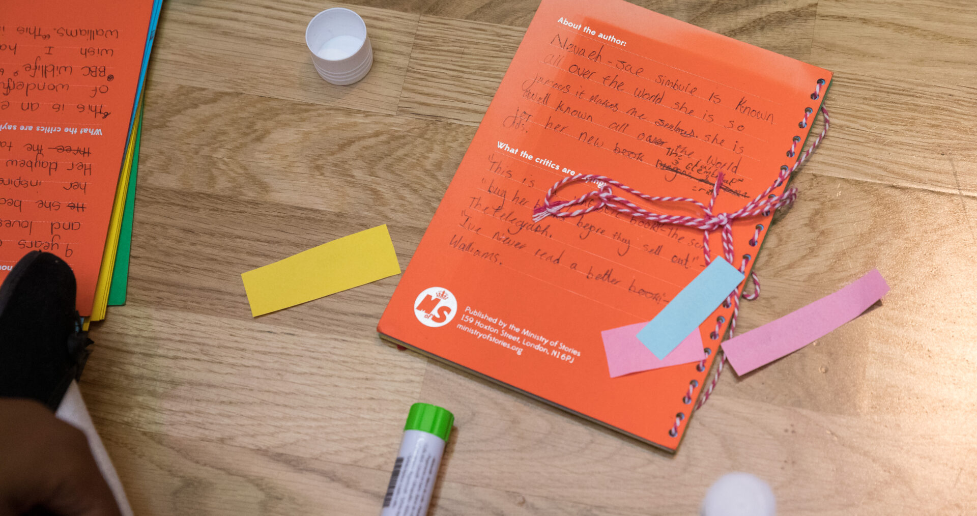 A orange book lays on a wooden floor. It is surrounding by colourful paper and glue sticks. The book is covered in a child's writing and looks inviting and fun.