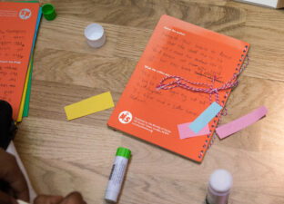 A orange book lays on a wooden floor. It is surrounding by colourful paper and glue sticks. The book is covered in a child's writing and looks inviting and fun.