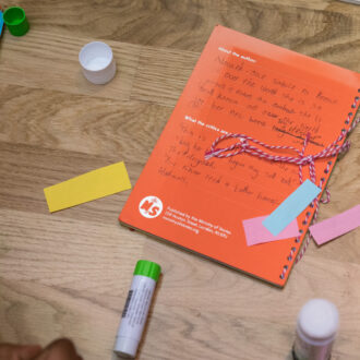 A orange book lays on a wooden floor. It is surrounding by colourful paper and glue sticks. The book is covered in a child's writing and looks inviting and fun.