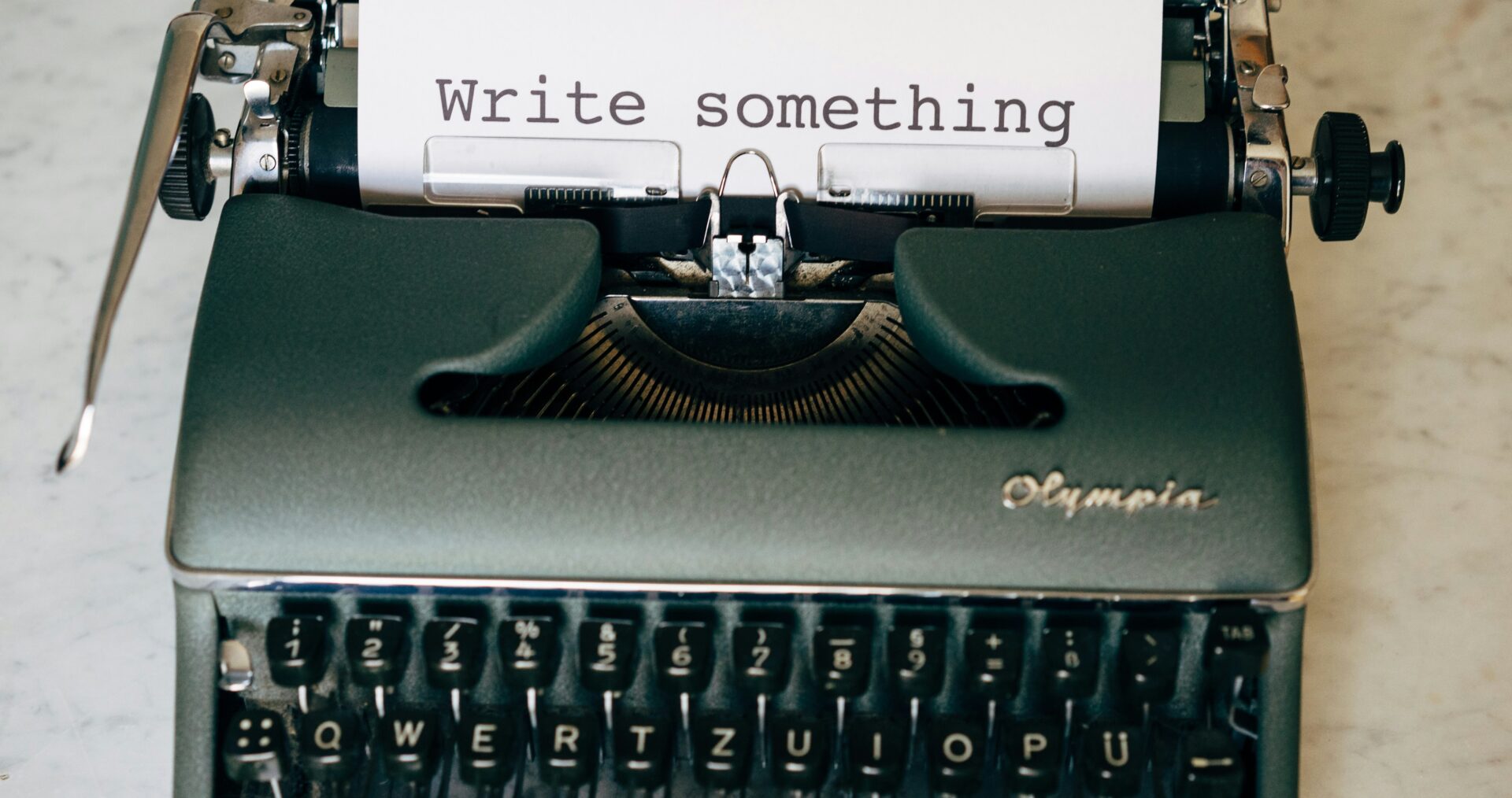 A grey typewriter sits on a cream, stone surface. A piece of water A4 paper, held in the typewriter, has the words typed 'write something'