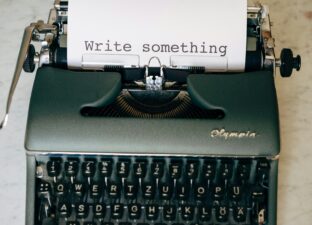 A grey typewriter sits on a cream, stone surface. A piece of water A4 paper, held in the typewriter, has the words typed 'write something'