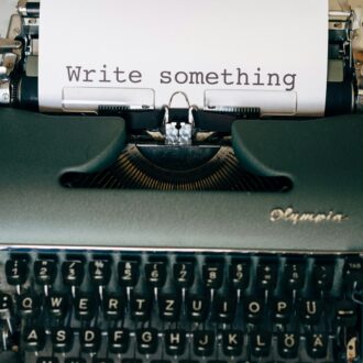 A grey typewriter sits on a cream, stone surface. A piece of water A4 paper, held in the typewriter, has the words typed 'write something'