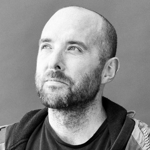 Paul Coomey sits looking into the distance. He has shaved head and short beard and wears a tshirt and hoody.