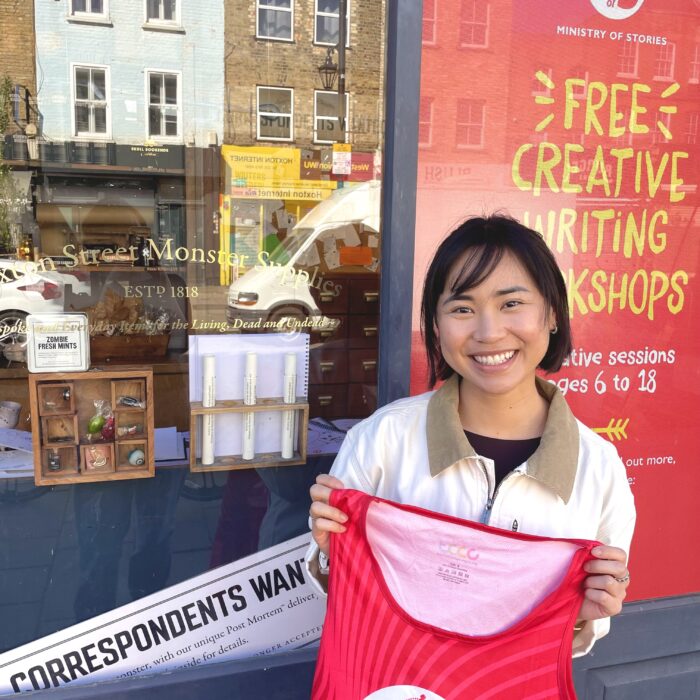 Tiffany, a woman with short dark hair, stands proudly in front of the Ministry of Stories building with her logoed runners vest