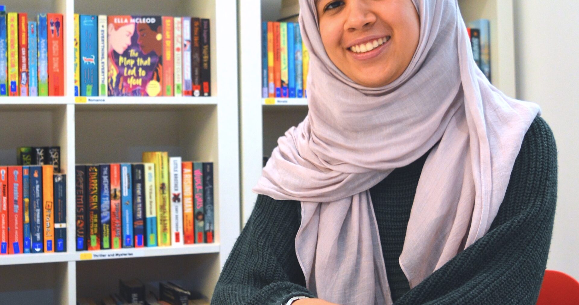 Fathima Zahra is a young Brown woman. She sits with her hands folded on the table in front of her and a huge, warm smile on her face. She is wearing a long sleeve dark blue top and a lilac hijab. Fathima has accessorised with a silver ring and silver bangle.