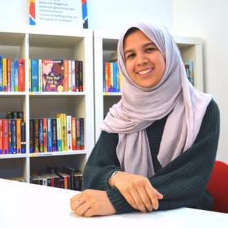 Fathima Zahra is a young Brown woman. She sits with her hands folded on the table in front of her and a huge, warm smile on her face. She is wearing a long sleeve dark blue top and a lilac hijab. Fathima has accessorised with a silver ring and silver bangle.