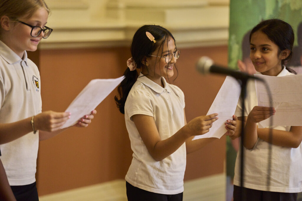 Three young girls stand smiling, holding speeches and addressing the audience with microphones