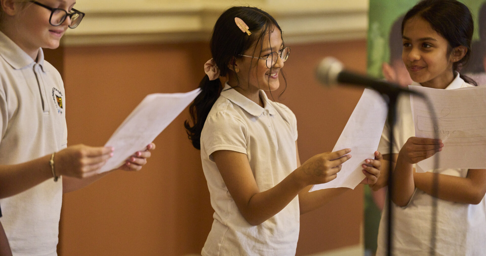 Three young girls stand smiling, holding speeches and addressing the audience with microphones