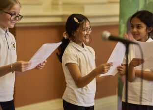 Three young girls stand smiling, holding speeches and addressing the audience with microphones