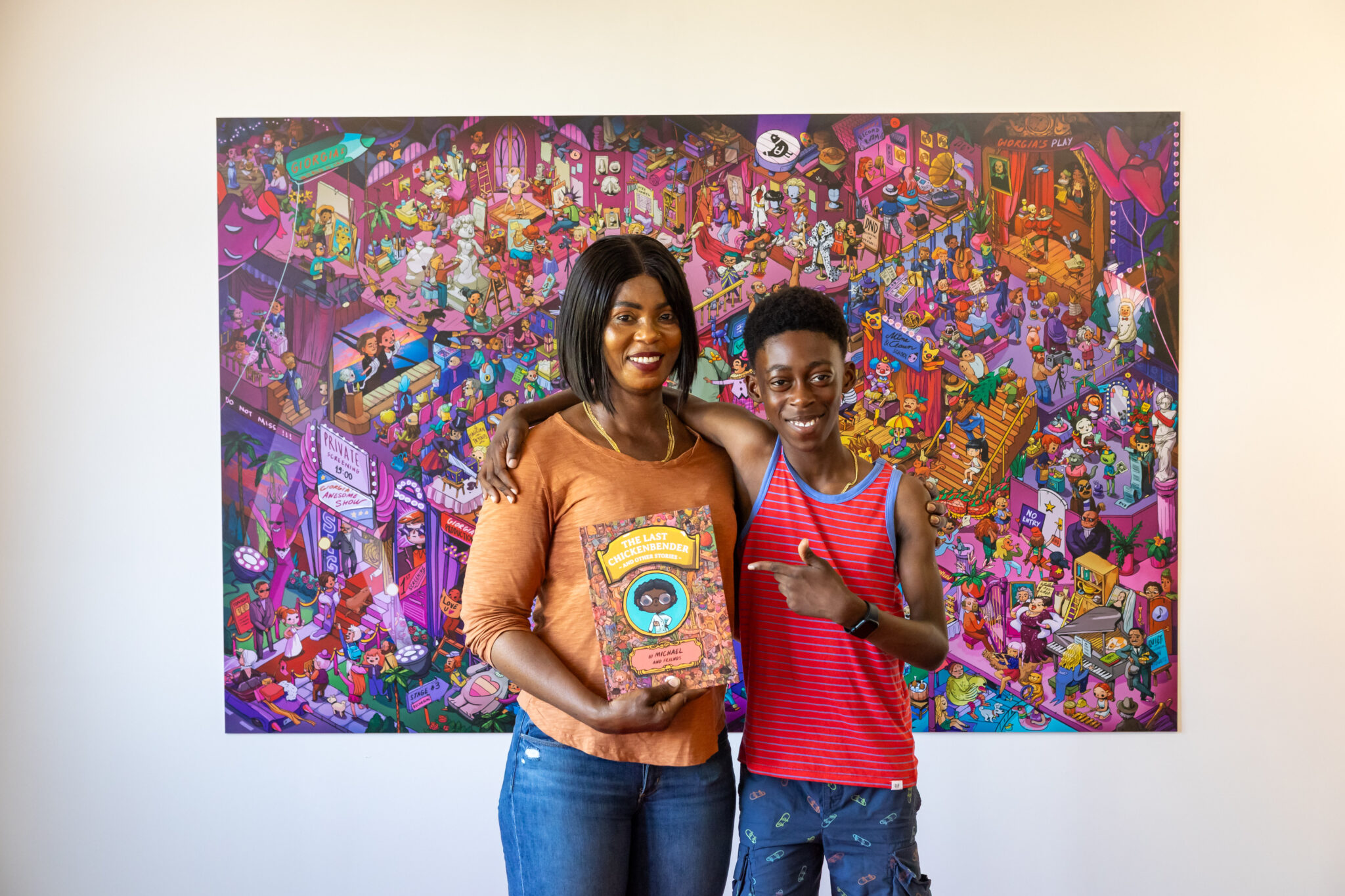 A young writer and his mum stand in front of a vibrant illustration of a Wonderbly world. She is clutching a book with his adventure story published inside and smiling. He has his arm around her and is pointing at the book.