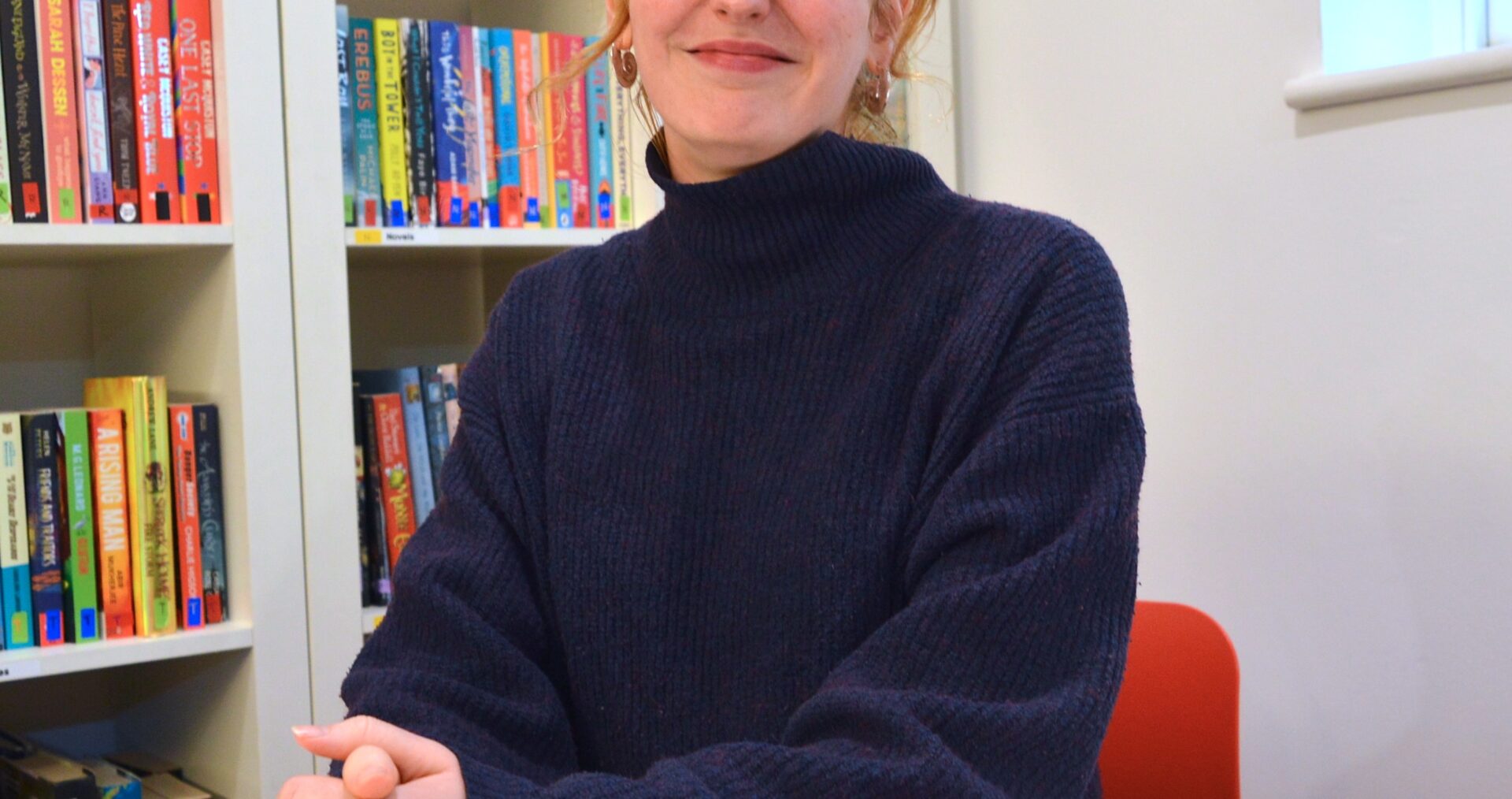 Lucie is a white woman, with blonde hair pulled back into a low ponytail with a full fringe. She wears a dark blue turtleneck jumper and smiles at the camera with her hands clasped in front of her.