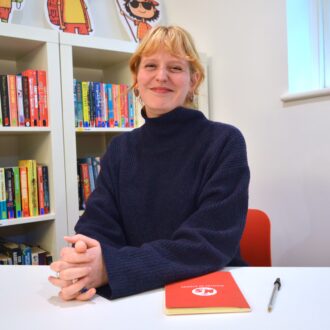 Lucie is a white woman, with blonde hair pulled back into a low ponytail with a full fringe. She wears a dark blue turtleneck jumper and smiles at the camera with her hands clasped in front of her.