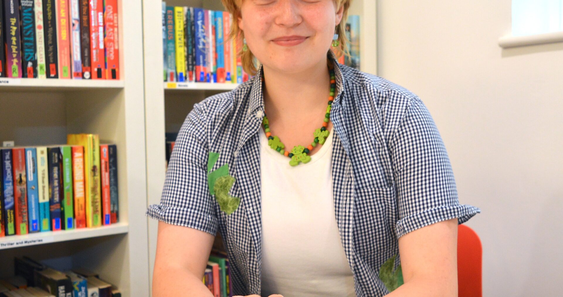 Nadia is a young white woman, with short dark blonde hair with a fringe. She wears a open, short sleeve cheque shirt with a white tshirt underneath and green beaded necklace. She clasps her hands in front of her and smiles to the camera.