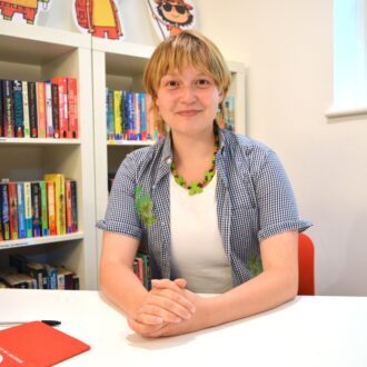 Nadia is a young white woman, with short dark blonde hair with a fringe. She wears a open, short sleeve cheque shirt with a white tshirt underneath and green beaded necklace. She clasps her hands in front of her and smiles to the camera.