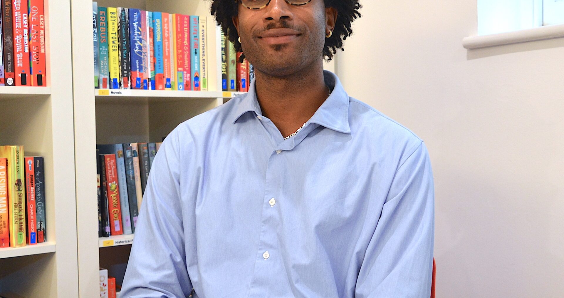 Rowan, a young black man with dark curly hair and glasses, wearing a blue open collar short, sits smiling at the camera with a red ministry of stories book in front of him .