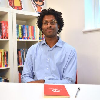 Rowan, a young black man with dark curly hair and glasses, wearing a blue open collar short, sits smiling at the camera with a red ministry of stories book in front of him .