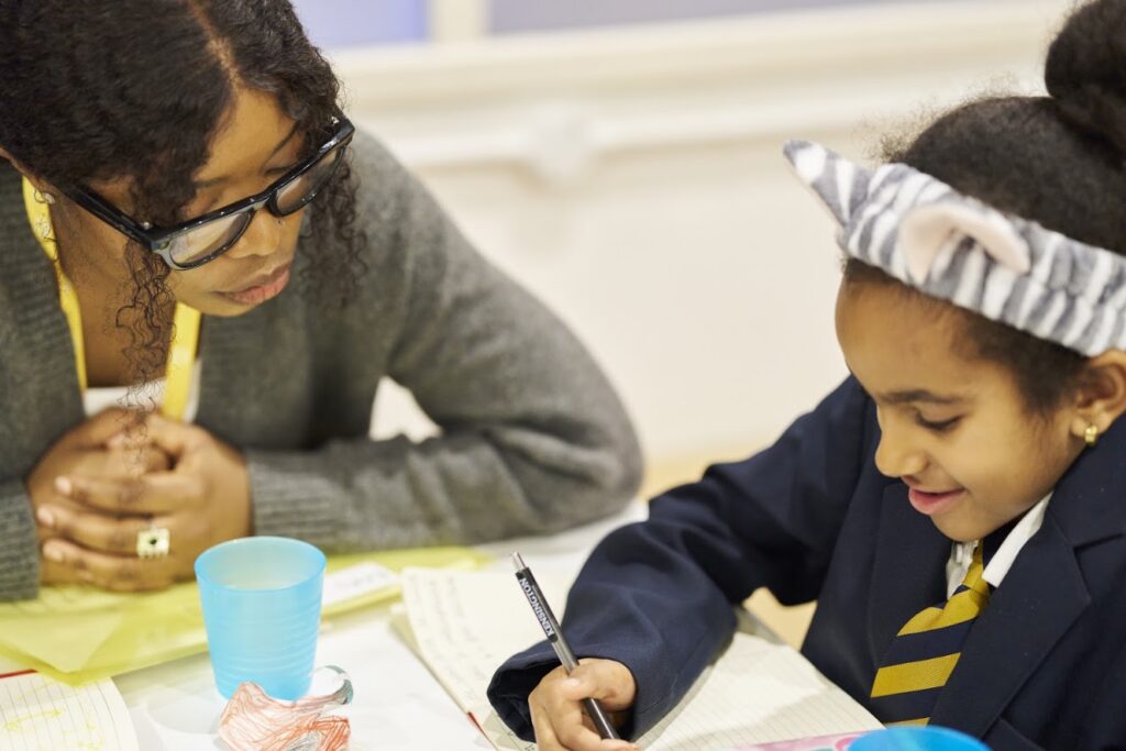 A mentor sits beside a young girl as she smiles and writes in her Ministry of Stories notebook