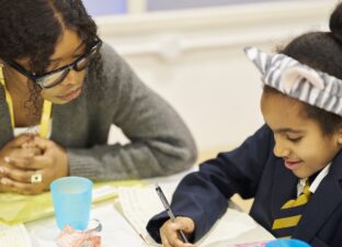 A mentor sits beside a young girl as she smiles and writes in her Ministry of Stories notebook